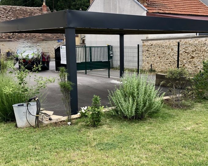 Carport moderne noir au-dessus d'une allée avec jardin verdoyant.